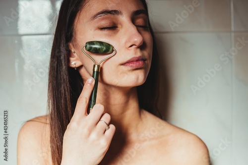 Crop concentrated young female with closed eyes massaging cheek with jade roller against ceramic wall photo