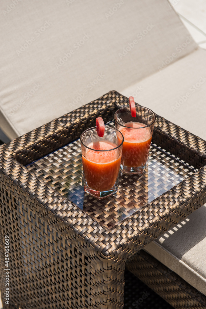 Glasses of fresh yummy watermelon squeezed juice served on small wicker rattan table near comfortable sunbed in tropical resort on sunny summer day Glasses of fresh yummy watermelon squeezed juice served on small wicker rattan table near comfortable sunbed in tropical resort on sunny summer day