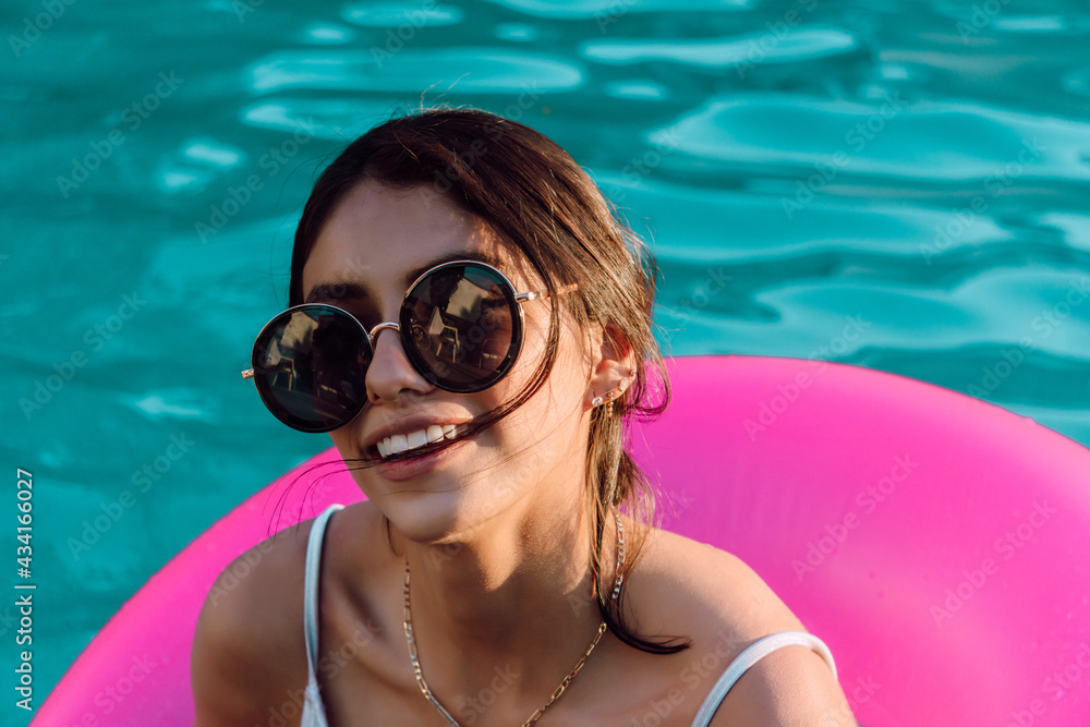 Young cheerful female traveler in swimwear and sunglasses lying in inflatable ring in swimming pool Young cheerful female traveler in swimwear and sunglasses lying in inflatable ring in swimming pool