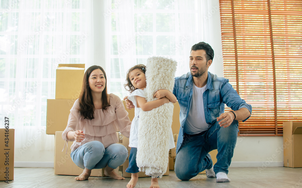 Little cute adorable curly hair daughter help her parents to carry carpet while moving house with background of father and mother and paper boxes. Insurance, Happiness and Real Estate Concept. Little cute adorable curly hair daughter help her parents to carry carpet while moving house with background of father and mother and paper boxes. Insurance, Happiness and Real Estate Concept.