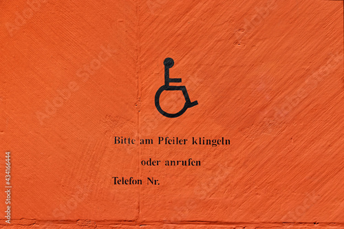 Handicapped symbol on an orange wall with german text, german text translation: please ring the bell at the post or call, phone number photo