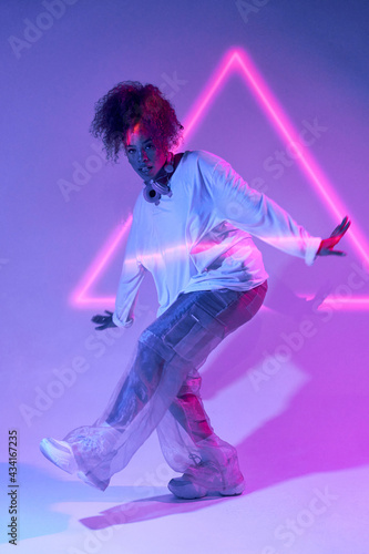 Full body talented young African American female dancer in loose clothes dancing in studio in neon lights and looking at camera photo