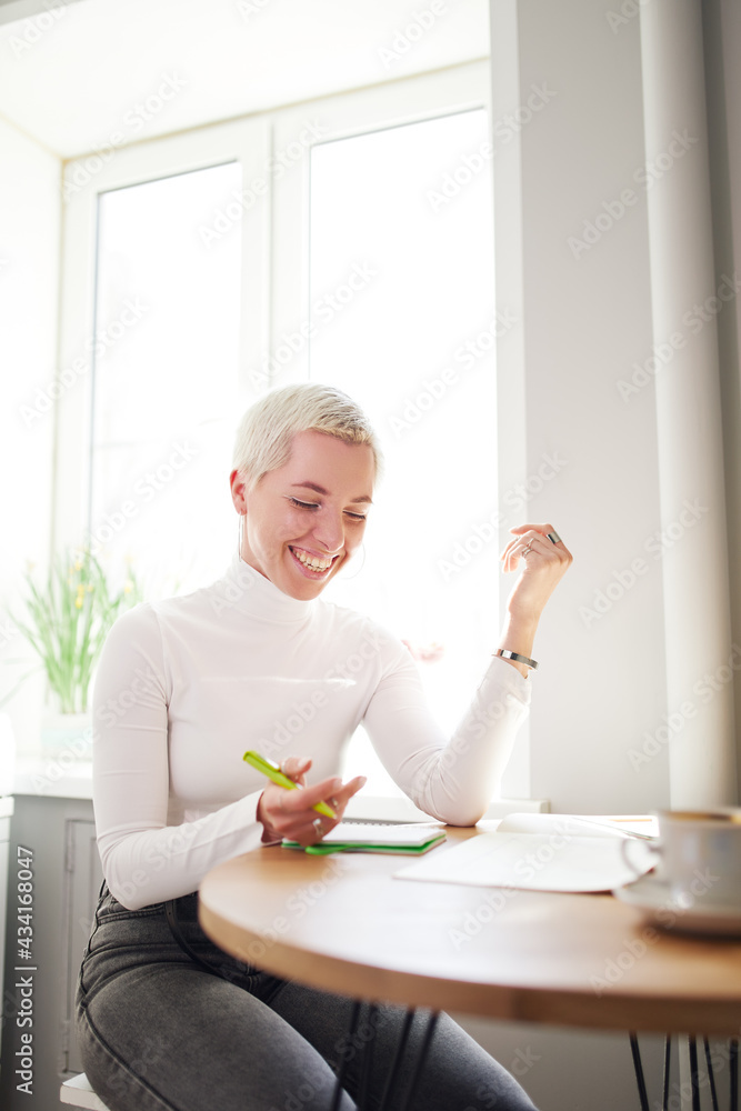 Side view of happy female astrologist taking notes in notepad at desk with cup of coffee at home in sunlight Side view of happy female astrologist taking notes in notepad at desk with cup of coffee at home in sunlight