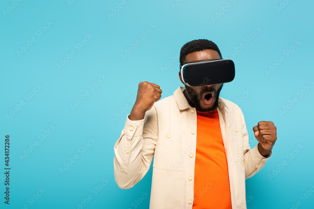 excited african american man in vr headset showing win gesture isolated on blue excited african american man in vr headset showing win gesture isolated on blue