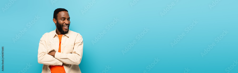 excited african american man with crossed arms laughing while looking away isolated on blue, banner excited african american man with crossed arms laughing while looking away isolated on blue, banner