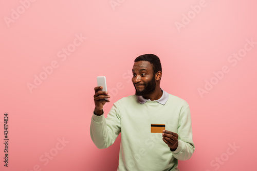 Smiling african american man using mobile phone and holding credit card on pink background