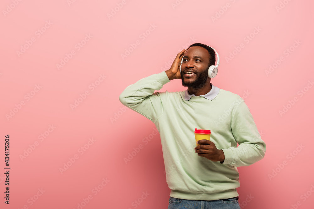 Smiling african american man holding paper cup and listening music in headphones on pink background Smiling african american man holding paper cup and listening music in headphones on pink background