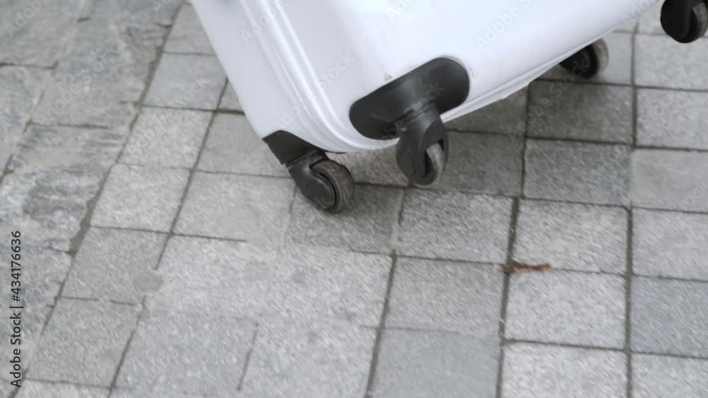 Black small wheels of white suitcase roll on gray paving slabs in the city. Close-up of travel bag Black small wheels of white suitcase roll on gray paving slabs in the city. Close-up of travel bag