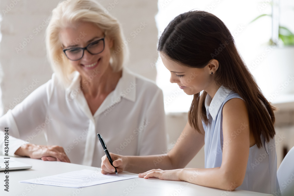 Smiling young Caucasian female employee put signature on contract close deal with middle-aged employer in office. Happy diverse businesswoman sign document make agreement after successful meeting. Smiling young Caucasian female employee put signature on contract close deal with middle-aged employer in office. Happy diverse businesswoman sign document make agreement after successful meeting.