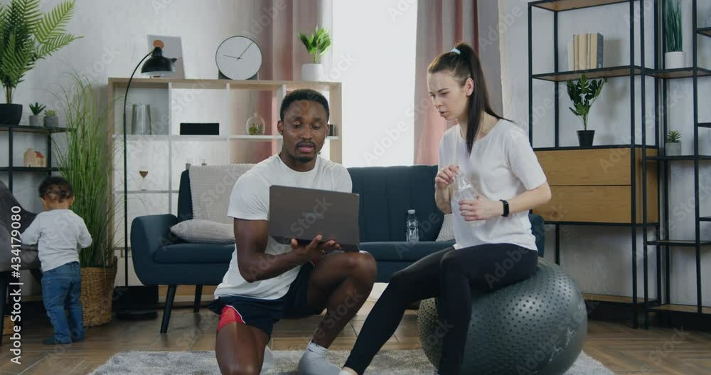Close up of good-looking positive modern young sporty mixed race couple which doing pause in joint workout ,man searching video training on laptop while woman sitting on fitball and drink water Close up of good-looking positive modern young sporty mixed race couple which doing pause in joint workout ,man searching video training on laptop while woman sitting on fitball and drink water