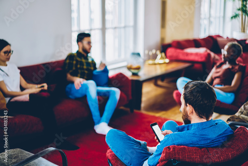 Back view of male blogger networking and chatting on blank smartphone during friendly meeting with blurred best friends for discussing text content on website, mock up screen on mobile phone