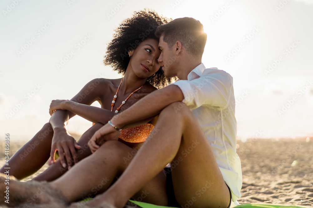 Interracial love couple tenderly touching the forehead sitting on the beach in the summertime