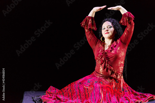 Gypsy woman barefoot with long black hair dances while sitting movements with her hands in a red dress on a black background.