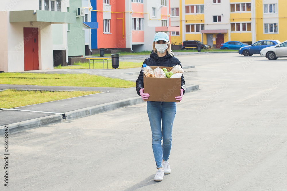 female volunteer wearing a protective mask walks down the street with a box of groceries, donations female volunteer wearing a protective mask walks down the street with a box of groceries, donations