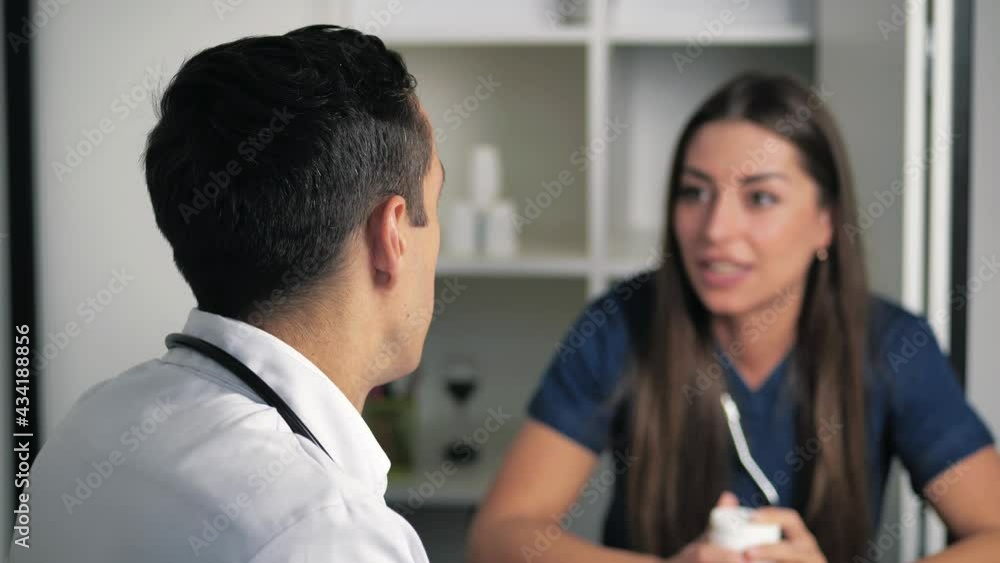Serious focused female physician in medical dark-blue coat sitting at table, consulting male doctor about illness or surgery. Rear view young man listening young doctor meeting. doctors consult Serious focused female physician in medical dark-blue coat sitting at table, consulting male doctor about illness or surgery. Rear view young man listening young doctor meeting. doctors consult