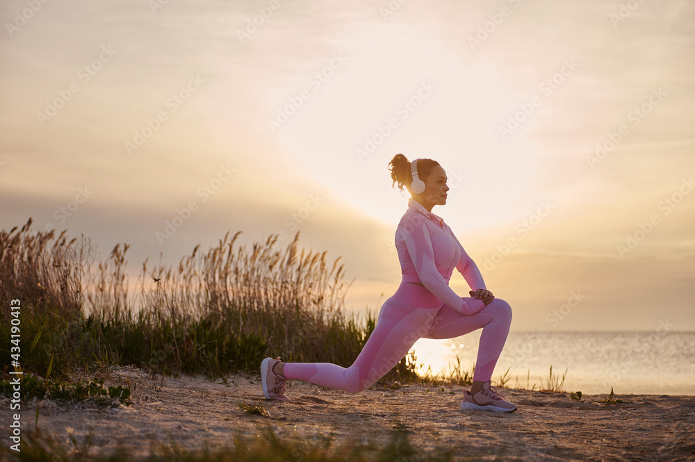 Young female athlete doing lunges and warming up at the estuary before an evening run at sunset Young female athlete doing lunges and warming up at the estuary before an evening run at sunset