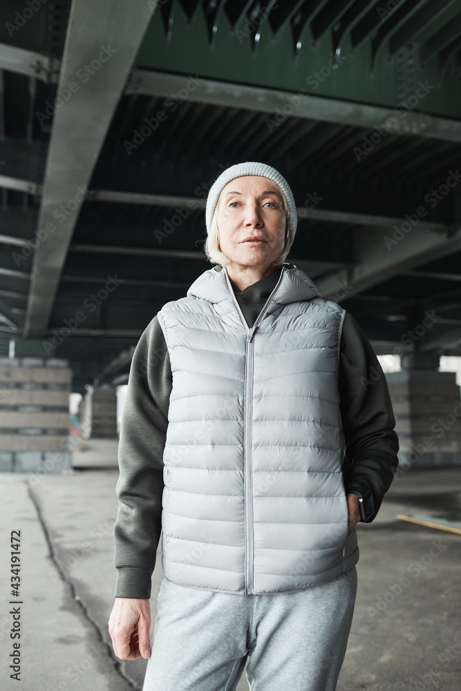 Portrait of serious sporty mature Caucasian woman in warm vest and hat standing confidently under bridge outdoors, gray color