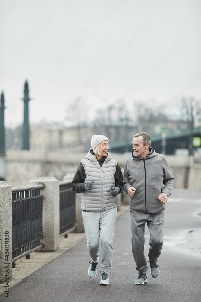 Happy active senior Caucasian couple in gray sport suits chatting and running along riverside while enjoying morning training