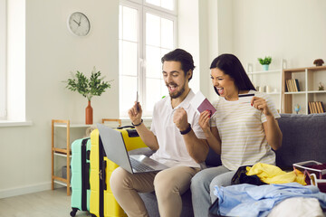 Happy couple getting ready for tour vacation trip pay for cheap hotel online. Excited boyfriend and girlfriend sitting on sofa with laptop, credit card, passports, flight tickets and travel suitcase