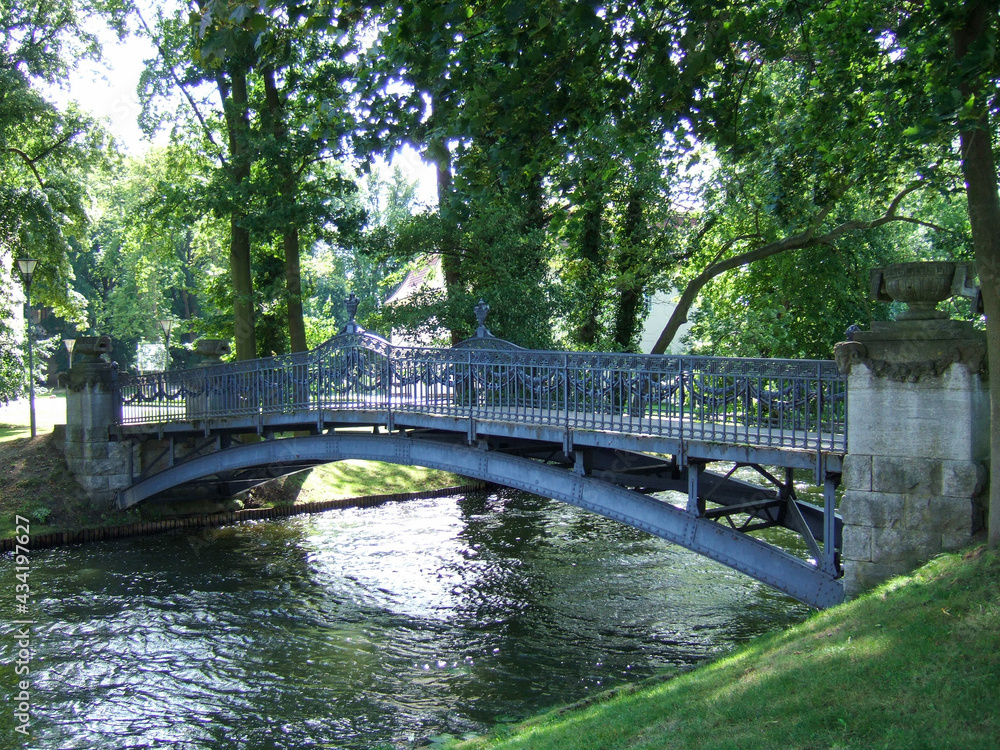 A curved, metal, deserted pedestrian bridge in a park over a river surrounded by green trees and meadows. The sun is reflected between the trees in the flowing water of the river. A curved, metal, deserted pedestrian bridge in a park over a river surrounded by green trees and meadows. The sun is reflected between the trees in the flowing water of the river.