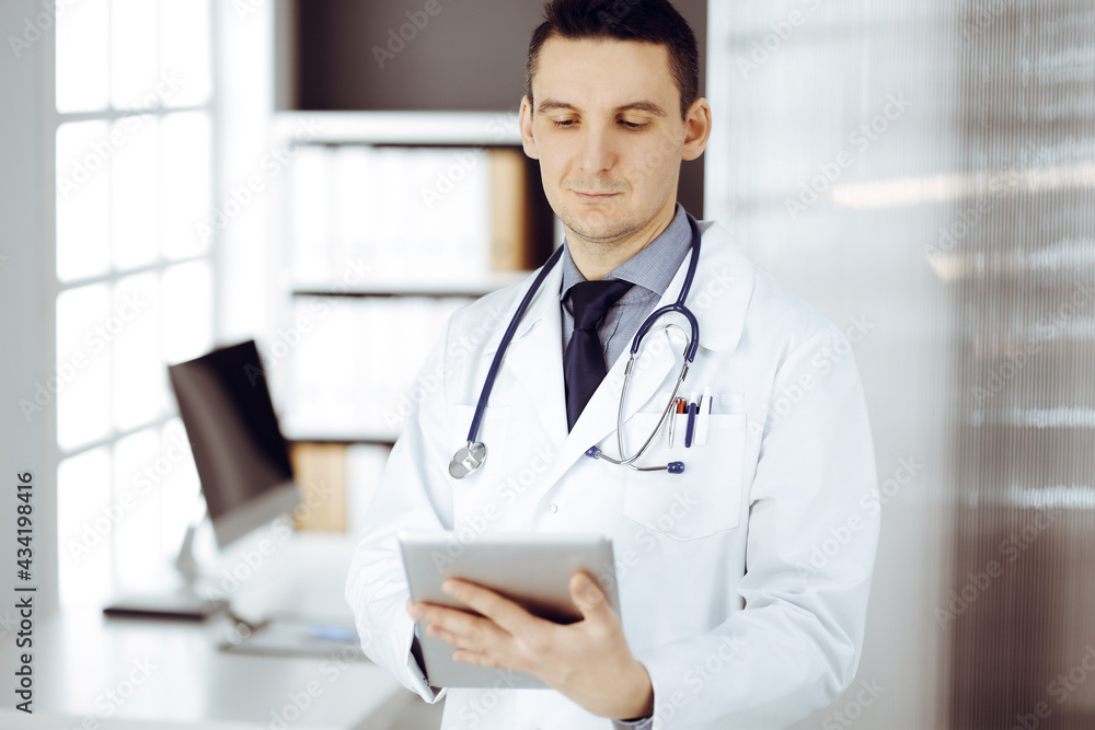 Friendly male doctor using tablet computer in sunny clinic. Medicine and healthcare concept Friendly male doctor using tablet computer in sunny clinic. Medicine and healthcare concept