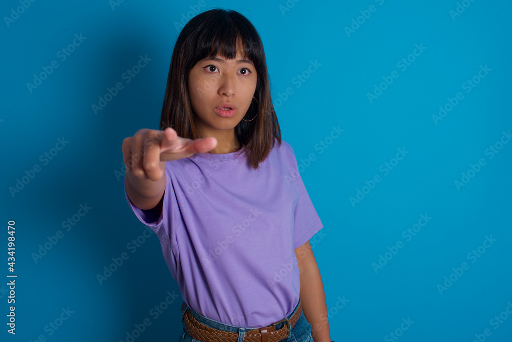 young beautiful asian woman wearing purple t-shirt against blue wall Pointing with finger surprised ahead, open mouth amazed expression, something on the front. young beautiful asian woman wearing purple t-shirt against blue wall Pointing with finger surprised ahead, open mouth amazed expression, something on the front.