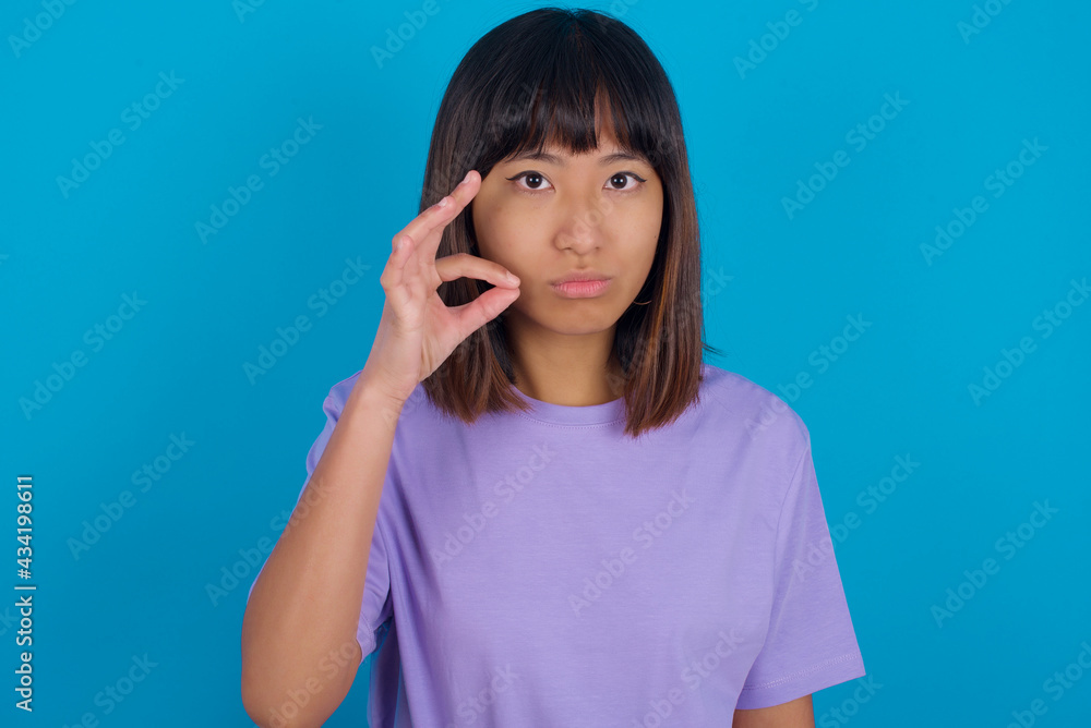 young beautiful asian woman wearing purple t-shirt against blue wall mouth and lips shut as zip with fingers. Secret and silent, taboo talking. young beautiful asian woman wearing purple t-shirt against blue wall mouth and lips shut as zip with fingers. Secret and silent, taboo talking.