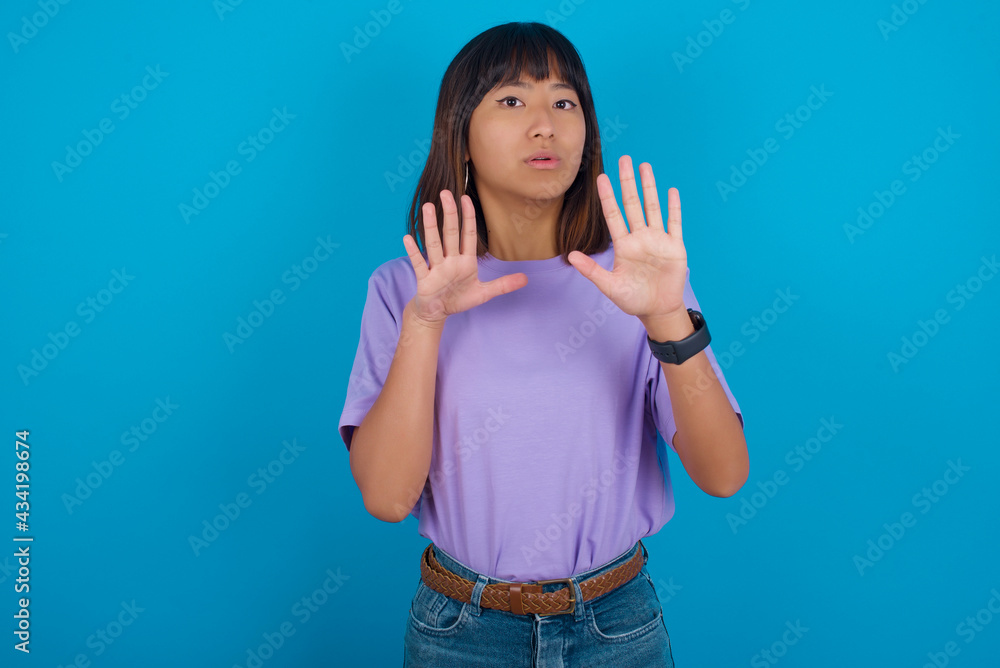 young beautiful asian woman wearing purple t-shirt against blue wall Moving away hands palms showing refusal and denial with afraid and disgusting expression. Stop and forbidden. young beautiful asian woman wearing purple t-shirt against blue wall Moving away hands palms showing refusal and denial with afraid and disgusting expression. Stop and forbidden.