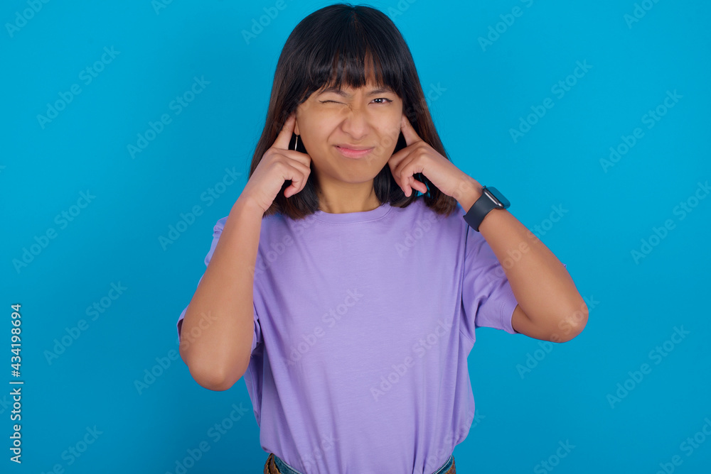 young beautiful asian woman wearing purple t-shirt against blue wall covering ears with fingers with annoyed expression for the noise of loud music. Deaf concept. young beautiful asian woman wearing purple t-shirt against blue wall covering ears with fingers with annoyed expression for the noise of loud music. Deaf concept.