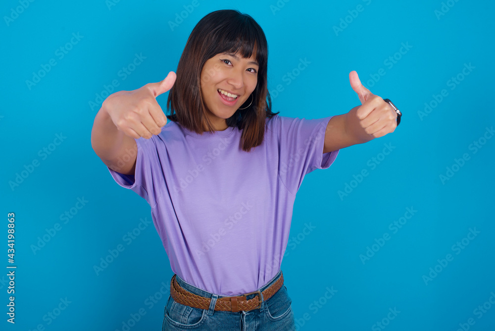 young beautiful asian woman wearing purple t-shirt against blue wall approving doing positive gesture with hand, thumbs up smiling and happy for success. Winner gesture. young beautiful asian woman wearing purple t-shirt against blue wall approving doing positive gesture with hand, thumbs up smiling and happy for success. Winner gesture.