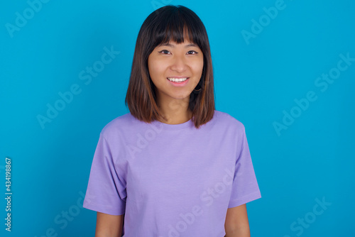 young beautiful asian woman wearing purple t-shirt against blue wall with a happy and cool smile on face. Lucky person.