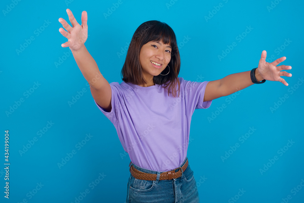 young beautiful asian woman wearing purple t-shirt against blue wall looking at the camera smiling with open arms for hug. Cheerful expression embracing happiness. young beautiful asian woman wearing purple t-shirt against blue wall looking at the camera smiling with open arms for hug. Cheerful expression embracing happiness.