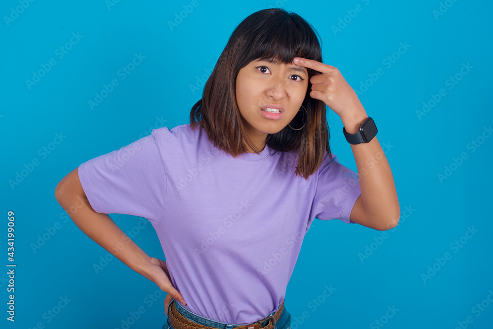 young beautiful asian woman wearing purple t-shirt against blue wall pointing unhappy at pimple on forehead, blackhead infection. Skincare concept. young beautiful asian woman wearing purple t-shirt against blue wall pointing unhappy at pimple on forehead, blackhead infection. Skincare concept.
