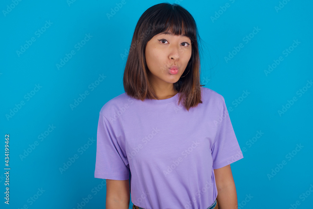 Shot of pleasant looking young beautiful asian woman wearing purple t-shirt against blue wall, pouts lips, looks at camera, Human facial expressions Shot of pleasant looking young beautiful asian woman wearing purple t-shirt against blue wall, pouts lips, looks at camera, Human facial expressions