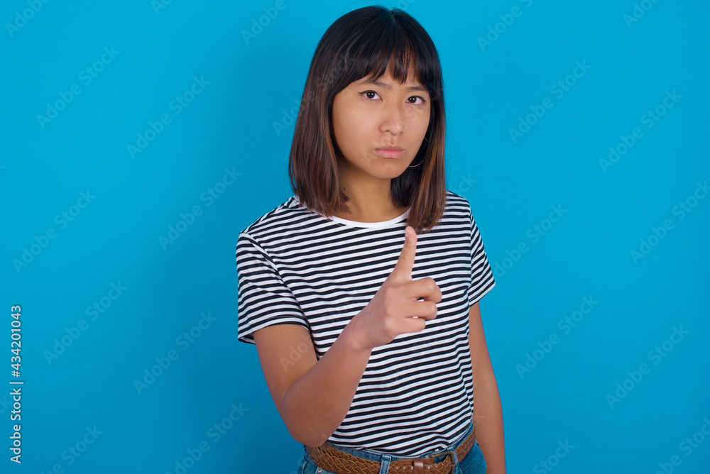 young beautiful asian woman wearing stripped t-shirt against blue wall frustrated and pointing to the front young beautiful asian woman wearing stripped t-shirt against blue wall frustrated and pointing to the front