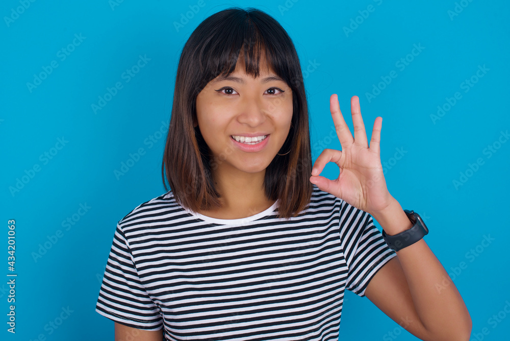 young beautiful asian woman wearing stripped t-shirt against blue wall hold hand arm okey symbol toothy approve advising novelty news young beautiful asian woman wearing stripped t-shirt against blue wall hold hand arm okey symbol toothy approve advising novelty news