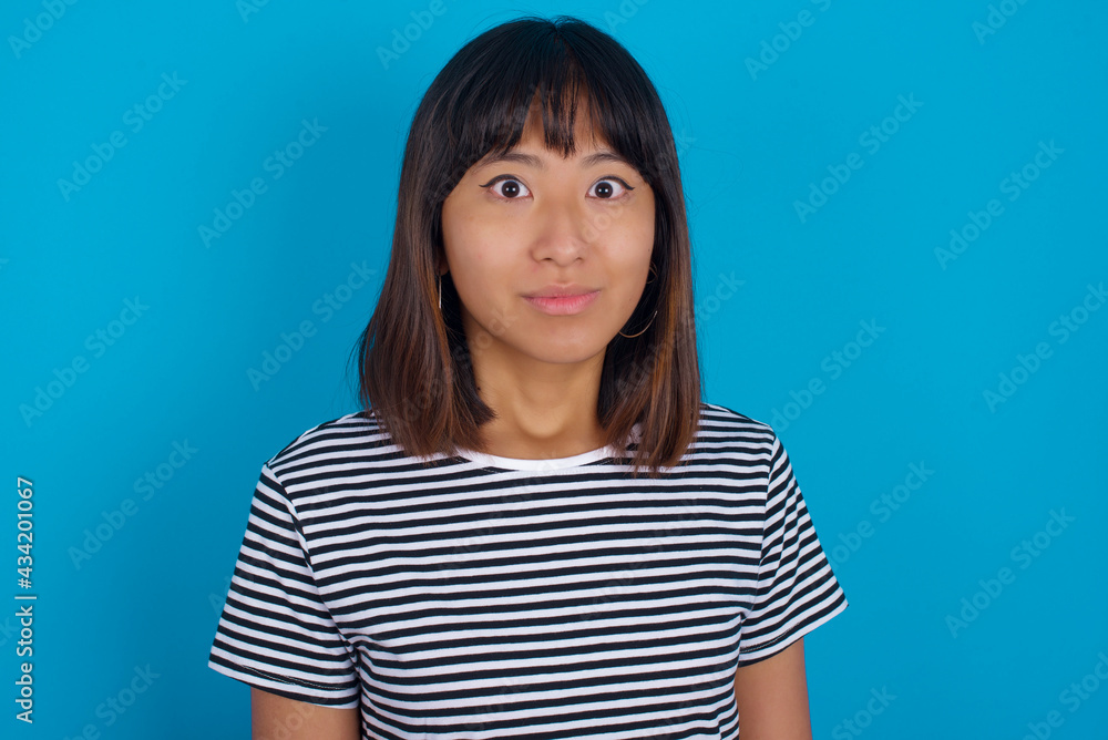 Stunned young beautiful asian woman wearing stripped t-shirt against blue wall stares reacts on shocking news. Astonished girl holds breath Stunned young beautiful asian woman wearing stripped t-shirt against blue wall stares reacts on shocking news. Astonished girl holds breath