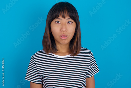 Shocked young beautiful asian woman wearing stripped t-shirt against blue wall stares bugged eyes keeps mouth opened has surprised expression. Omg concept