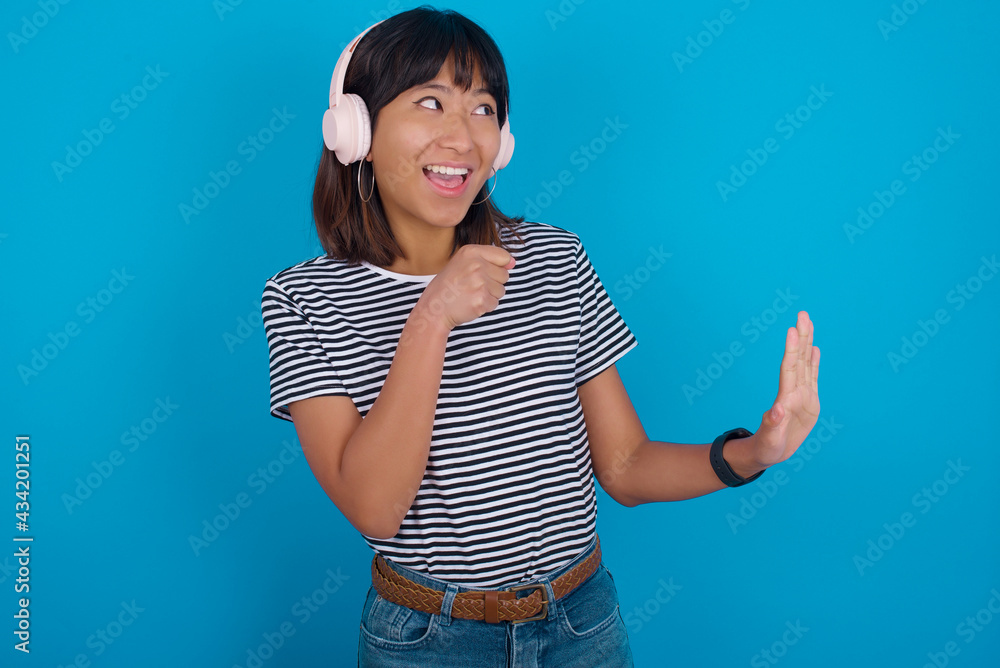 Fototapeta premium Happy young beautiful asian woman wearing stripped t-shirt against blue wall sings favourite song keeps hand near mouth as if microphone wears wireless headphones, listens music