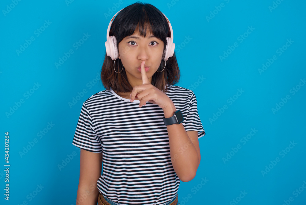 young beautiful asian woman wearing stripped t-shirt against blue wall making hush gesture with finger on her lips wearing wireless headphones. Be quiet. young beautiful asian woman wearing stripped t-shirt against blue wall making hush gesture with finger on her lips wearing wireless headphones. Be quiet.