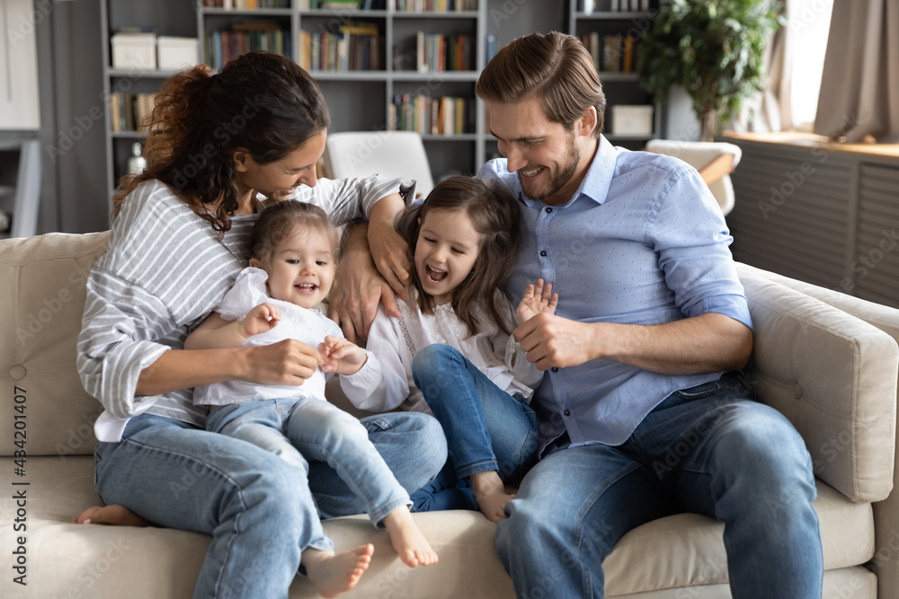 Overjoyed young Caucasian family with two small daughters sit relax on sofa have fun playing game together on weekend. Happy parents with little girls children kids feel playful tickle and laugh.