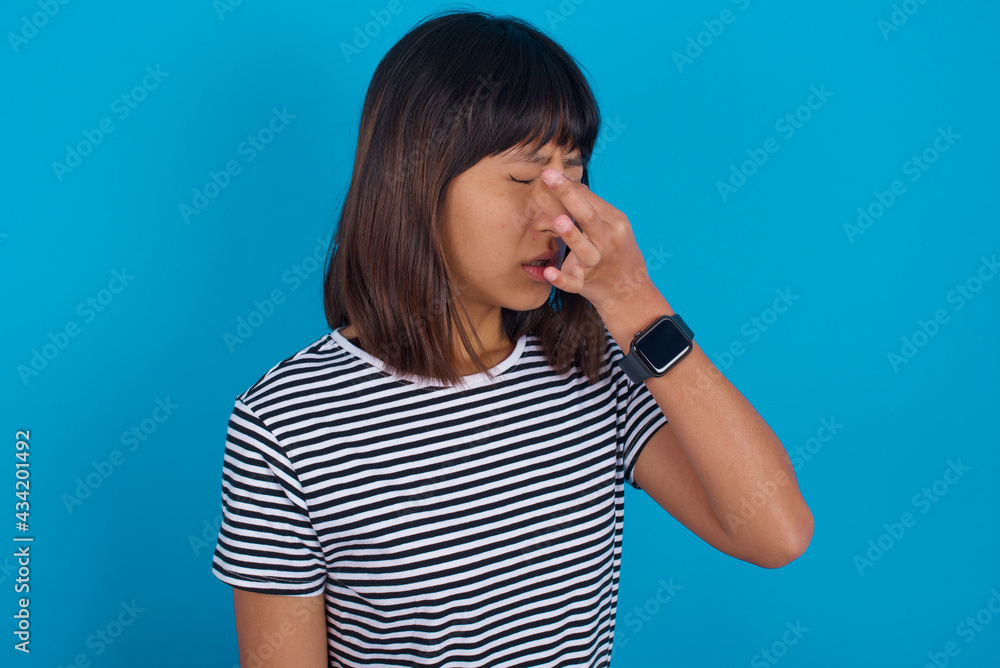 Sad young beautiful asian woman wearing stripped t-shirt against blue wall suffering from headache holding hand on her face Sad young beautiful asian woman wearing stripped t-shirt against blue wall suffering from headache holding hand on her face