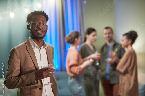 Waist up portrait of elegant African-American man looking at camera and holding cocktail glass while enjoying party indoors, copy space