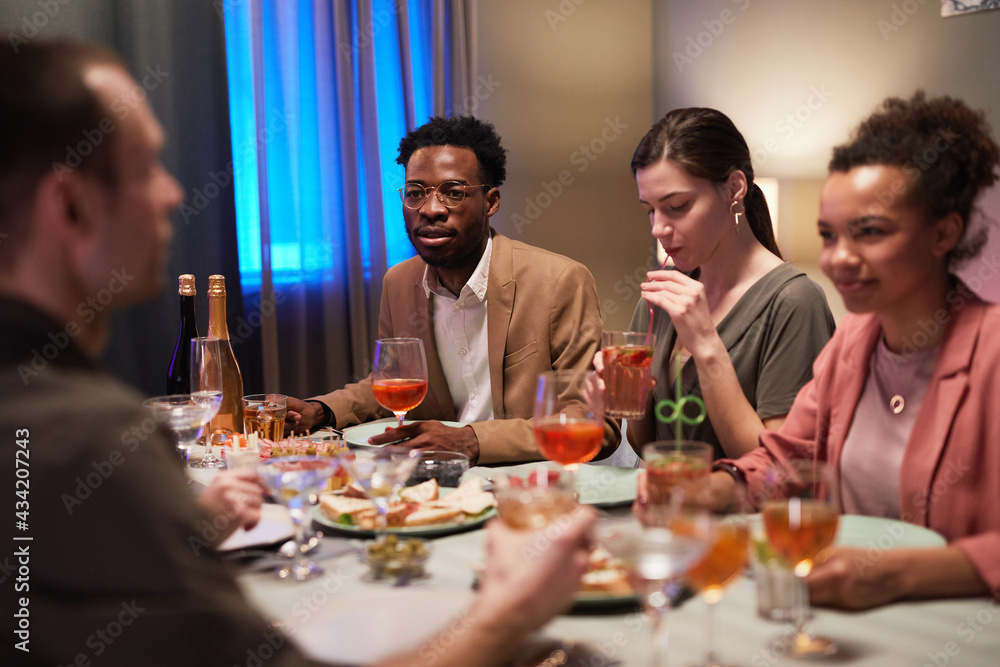 Diverse group of young people sitting at dinner table while enjoying party indoors Diverse group of young people sitting at dinner table while enjoying party indoors