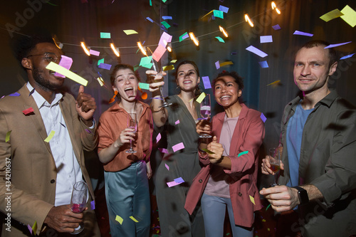 Multi-ethnic group of carefree people dancing in confetti while enjoying party indoors, shot with flash