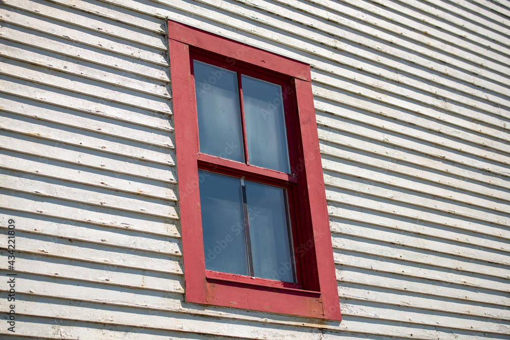 A bright red double hung vintage window with four glass panes. The residential window is on the exterior of a narrow horizontal white wooden clapboard siding wall. The clouds are reflecting in window.