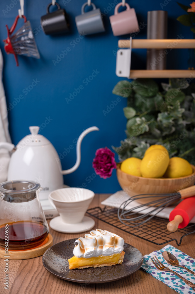 Lemon meringue pie in a black plate on a wooden counter with black coffee, teapot, server, lemon, and baking utensils as a decoration.