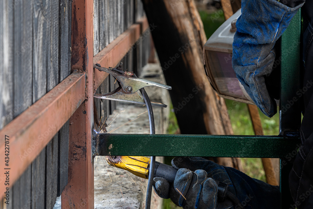 Close up of the a welder welds a fence on the construction site in the village. Construction on a metal frame. Close up of the a welder welds a fence on the construction site in the village. Construction on a metal frame.