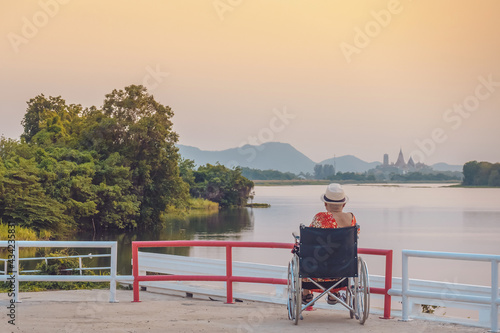Back view of Asian elderly female patient wearing surgical mask and sitting on wheelchair with feeling lonely and depressed at riverside in the evening with view of Wat Tham Sua in background.