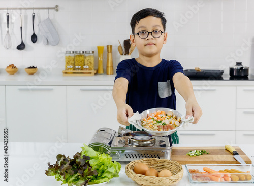 Kid using white cloths to carefully pick up hot flying pan of salad, hotdogs, hams, and crab stick out from gas stove in kitchen and show it to camera. photo